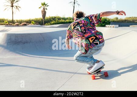 Vue arrière de l'anonyme funky jeune homme skateboarder dans le mode chemise et jean colorés en train de jouer sur une rampe en béton compétences de pratique dans le parc de skate Banque D'Images