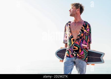 Jeune boxer taille basse pour homme avec une tenue tendance pour l'été portant un skateboard tout en se tenant sur un remblai contre la mer et un ciel sans nuages dedans jour d'été Banque D'Images