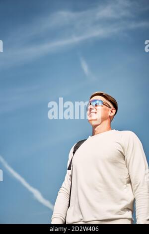 Du dessous du corps entier de jeunes hommes confiants dans le mode tenue décontractée et lunettes de soleil avec sac sur l'épaule marchant le long rue urbaine avec bâtiments contemporains en arrière-plan Banque D'Images