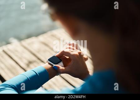 Vue arrière d'une athlète féminine inreconnaissable assise sur tapis et impulsion de contrôle sur le tracker d'entraînement en se reposant après entraînement en plein air Banque D'Images