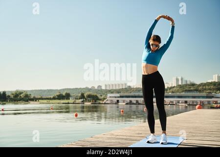Corps complet de jeune femme en vêtements de sport et lunettes de soleil étirez les bras tout en vous réchauffant pendant votre entraînement physique sur du bois jetée près de la rivière Banque D'Images