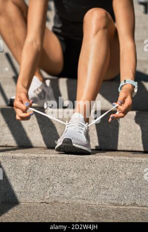 Faible angle de la récolte athlète anonyme féminine assis sur la pierre escaliers et lacets de chaussures sur les baskets lors de la préparation pour l'extérieur formation Banque D'Images