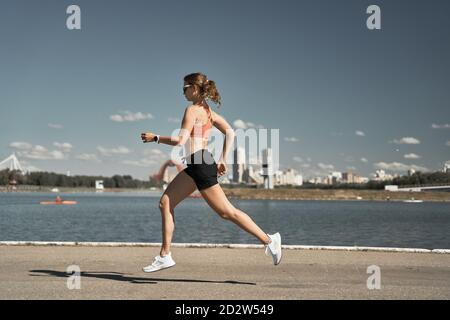 Vue latérale de l'ensemble du corps de la femme dynamique et motivée activewear avec tracker de forme physique jogging sur le remblai pavé près de la rivière en ville Banque D'Images