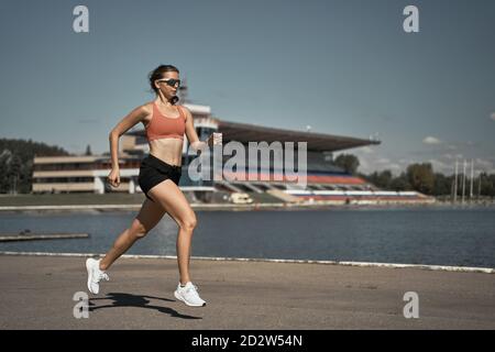 Vue latérale de l'ensemble du corps de la femme dynamique et motivée activewear avec tracker de forme physique jogging sur le remblai pavé près de la rivière en ville Banque D'Images