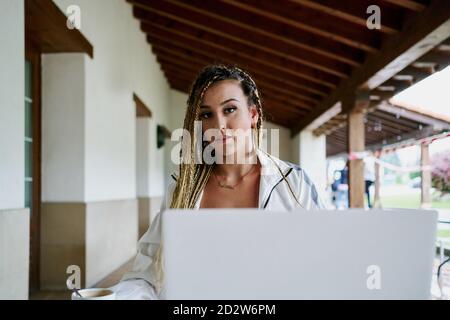 Femme entrepreneur avec des tresses élégantes assis à une table avec un ordinateur portable et travailler à distance dans un café tout en regardant la caméra Banque D'Images