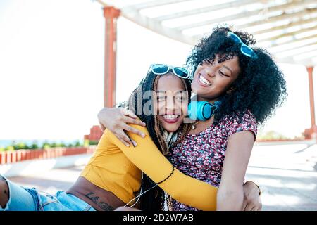 Des amies noires gaies avec des tresses et des cheveux bouclés assis se serrer les uns les autres sur les berges et s'amuser en été Banque D'Images