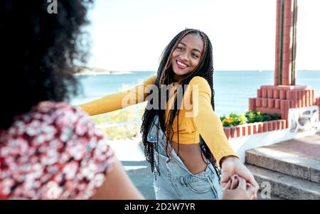 Ethnique curly court anonyme femme avec des cheveux bouclés tenant la main de la femme avec des tresses tout en se tenant debout sur la promenade en été et regardant l'un l'autre Banque D'Images