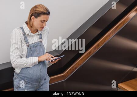 Femme enceinte concentrée dans une combinaison en denim près de l'escalier Bâtiment contemporain et accès Internet par téléphone portable Banque D'Images