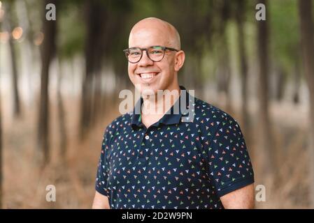 Homme beau positif avec un sourire en dents de souris et dans des lunettes debout dans une allée avec des arbres et regardant la caméra Banque D'Images