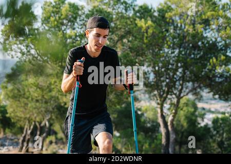 Jeune homme sportif positif en randonnée active avec bâtons de randonnée sur un sentier pierreux dans un terrain montagneux verdoyant et boisé Banque D'Images