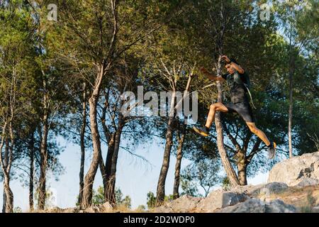 Vue du dessous du côté actif de la sportive homme saut haut sur un sentier rocheux tout en courant dans une forêt verte en montagne Banque D'Images
