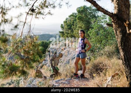 Corps complet de la jeune femme en vêtements actifs debout sur la pierreuse sentier tout en prenant le repos pendant la course sur sentier dans les hautes terres boisées en été Banque D'Images