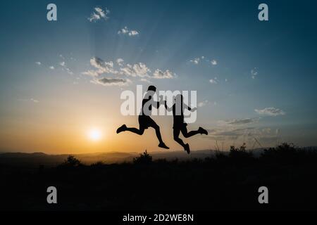 Silhouettes avec vue latérale d'amis masculins et féminins qui sautent haut au-dessus du sol tout en s'amusant et en profitant de la liberté terrain vallonné contre le coucher du soleil Banque D'Images