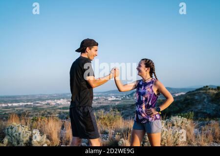 Vue latérale d'un jeune homme et d'une femme heureux en mode actif se saluant les uns les autres avec un mouvement des bras de lutte tout en préparant la course à pied ensemble en terrain montagneux Banque D'Images