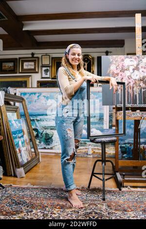 Ensemble de jeunes femmes artistiques créatives en denim tenir le cadre vide tout en se tenant près du chevalet avec l'image à l'intérieur atelier de peintures Banque D'Images