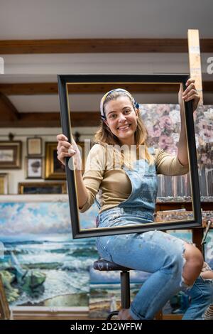 Jeune artiste créative en denim ensemble tenant un cadre vide tout en étant assis près du chevalet avec une photo dans un atelier avec des peintures Banque D'Images