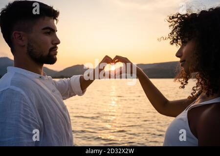 Couple de race mixte, homme caucasien avec sa petite amie hispanique, formant un cœur avec leurs mains encadrant le soleil couchant sur un lac d'eau - multiethnique Banque D'Images