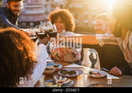 Les jeunes gens multiraciaux applaudissent avec du vin lors du dîner de fête pendant Porter des masques de protection - concept de distance sociale - Focus on mains lunettes Banque D'Images