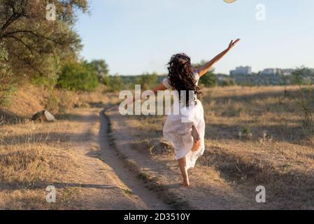 une fille heureuse court dans le champ le long du chemin, rebondissant et agitant ses bras. Sur fond de soleil d'été. Banque D'Images