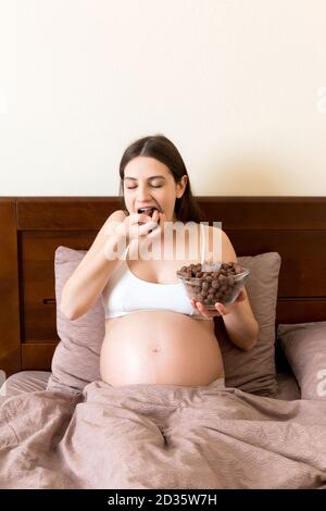 La femme enceinte mange des boules de céréales croquantes au chocolat dans un bol qui se repose au lit. Profiter de la nourriture pendant la grossesse concept. Banque D'Images