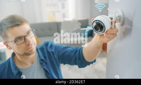 Jeune homme en lunettes portant un maillot bleu est en train de régler une caméra de surveillance Wi-Fi moderne avec deux antennes sur un mur blanc à la maison. Banque D'Images