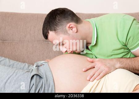 Jeune homme barbu mettant son oreille près de son abdomen de femme enceinte et l'embrassant. Banque D'Images