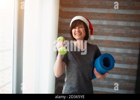 La femme fine pour adulte s'entraîner à la maison. Personne féminine senior adulte gaie pendant la période de noël ou du nouvel an. Tenue d'une haltère et yoga Banque D'Images