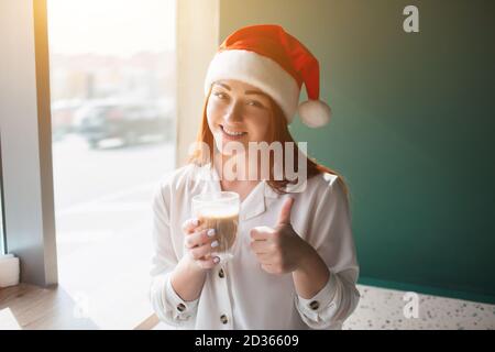 Le modèle féminin en chapeau de noël montre le pouce vers le haut. Une jeune femme boit du café et sourit Banque D'Images