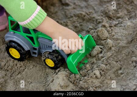 partie de l'image d'un petit enfant qui s'assoit dans le sable humide et joue avec un véhicule de construction de jouet vert. Main visible, vêtue d'un blou vert Banque D'Images