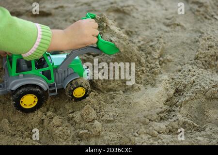 partie de l'image d'un petit enfant qui s'assoit dans le sable humide et joue avec un véhicule de construction de jouet vert. Main visible, vêtue d'un blou vert Banque D'Images