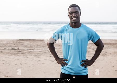 Joyeux athlète noir tenant les mains à la taille et souriant pour l'appareil photo tout en se tenant sur la plage de sable Famara près de la mer à Lanzarote, Espagne Banque D'Images