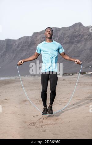 Athlète afro-américain de pleine longueur sautant avec une corde sur la plage de sable de Famara pendant l'entraînement de remise en forme à Lanzarote, Espagne Banque D'Images