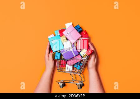 Panier de Noël plein de cadeaux colorés, isolé sur fond orange. Vue de dessus avec une femme achetant des cadeaux. Achats en ligne pour des cadeaux de Noël. Banque D'Images