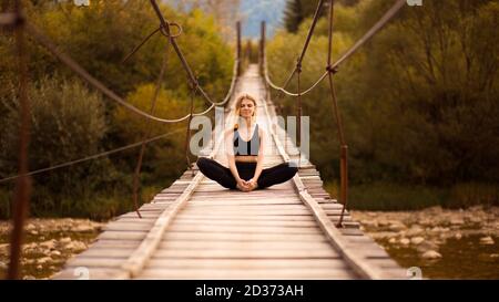 Pratique respiratoire. La jeune femme en forme est assise dans la posture du papillon de yoga sur le pont au-dessus de la rivière de montagne. La femme fait de l'exercice d'étirement de yoga ou de médit Banque D'Images