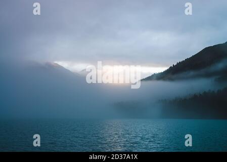 Ondule sur la surface du lac de montagne dans la brume en heure d'or. Le soleil brille à travers des nuages denses bas dans les montagnes. Le sentier ensoleillé brille sur l'eau au lever du soleil. A Banque D'Images