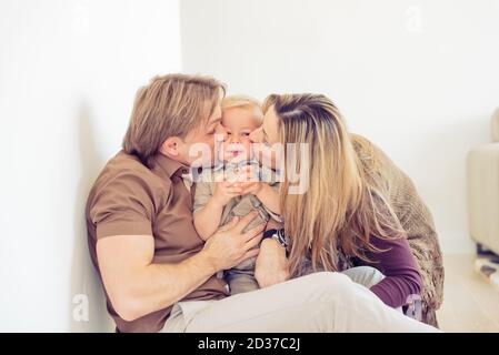 Famille heureuse assise sur le sol avec leur petit bébé. La famille passe du temps à la maison avec son fils. Parents embrassant leur enfant. Banque D'Images