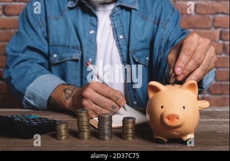 Hipster homme mettant la pièce dans la banque de porc avec pile de pièces sur la table pour économiser de l'argent. Banque D'Images