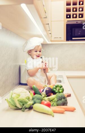 Bébé garçon assis parmi les légumes dans la cuisine et prépare pour cuisiner Banque D'Images