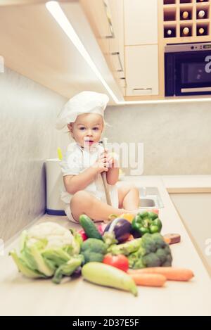 Bébé garçon assis parmi les légumes dans la cuisine et prépare pour cuisiner Banque D'Images