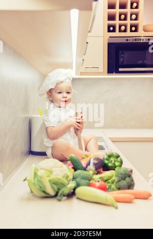 Bébé garçon assis parmi les légumes dans la cuisine et prépare pour cuisiner Banque D'Images