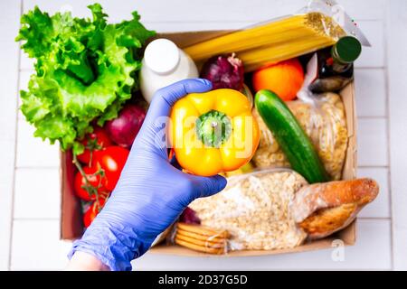 Faites du bénévolat en portant des gants pour donner de la nourriture dans une boîte en carton contenant divers aliments. Ouvrir la boîte en carton contenant de l'huile, des légumes, du lait, des conserves, des céréales et Banque D'Images