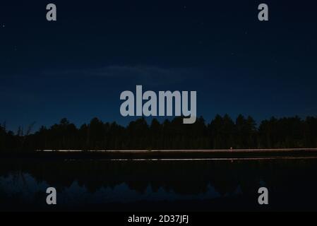 Réflexion de sentiers de lumière d'un véhicule sur un petit lac la nuit, placé contre les arbres et le ciel de nuit. Banque D'Images