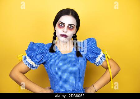 femme portant une poupée effrayante déguisement d'halloween sur fond jaune sceptique et nerveux, désapprouvant l'expression sur le visage avec les bras à la taille Banque D'Images