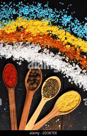 Concept de l'art alimentaire. Composition de sel bleu, blanc, orange et jaune. Cuillères en bois avec paprika, herbes, curcuma et boules de poivre près de sel de mer granulé. Ensemble d'épices sur fond de bois gris foncé. Banque D'Images