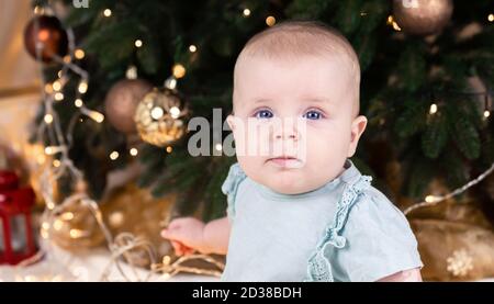 Bébé de Noël regardant l'appareil photo. Une petite fille mignonne dans une robe bleue et un chapeau blanc exprime ses émotions. Concept de Noël avec petit enfant, arbre et Banque D'Images