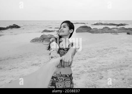 Jeune heureuse belle femme asiatique souriant et pensant tout en tenant mains et homme dirigeant à la plage Banque D'Images