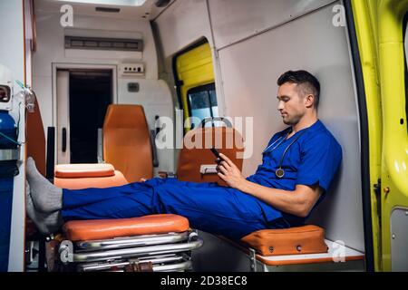 Jeune stagiaire dans un uniforme bleu se reposant et parcourant son téléphone dans la voiture d'ambulance. Banque D'Images
