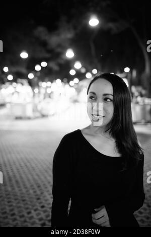 Young happy Asian woman smiling et penser contre vue panoramique sur le marché de nuit de Hua Hin, Thaïlande Banque D'Images