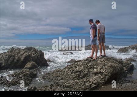 Deux jeunes hommes debout sur les rochers regardant le surf Banque D'Images