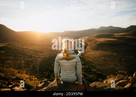 Jeune adolescente assise sur un rocher surplombant la terre verte au coucher du soleil après une randonnée réussie. Banque D'Images